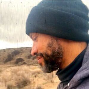 picture of the side of tyrone martinez black's face. Ty is black, and is wearing a beanie and what looks like a loose turtleneck shirt, they have a beard and mustache and are smiling. In the background you see an outdoor landscape of brownish and tan land.